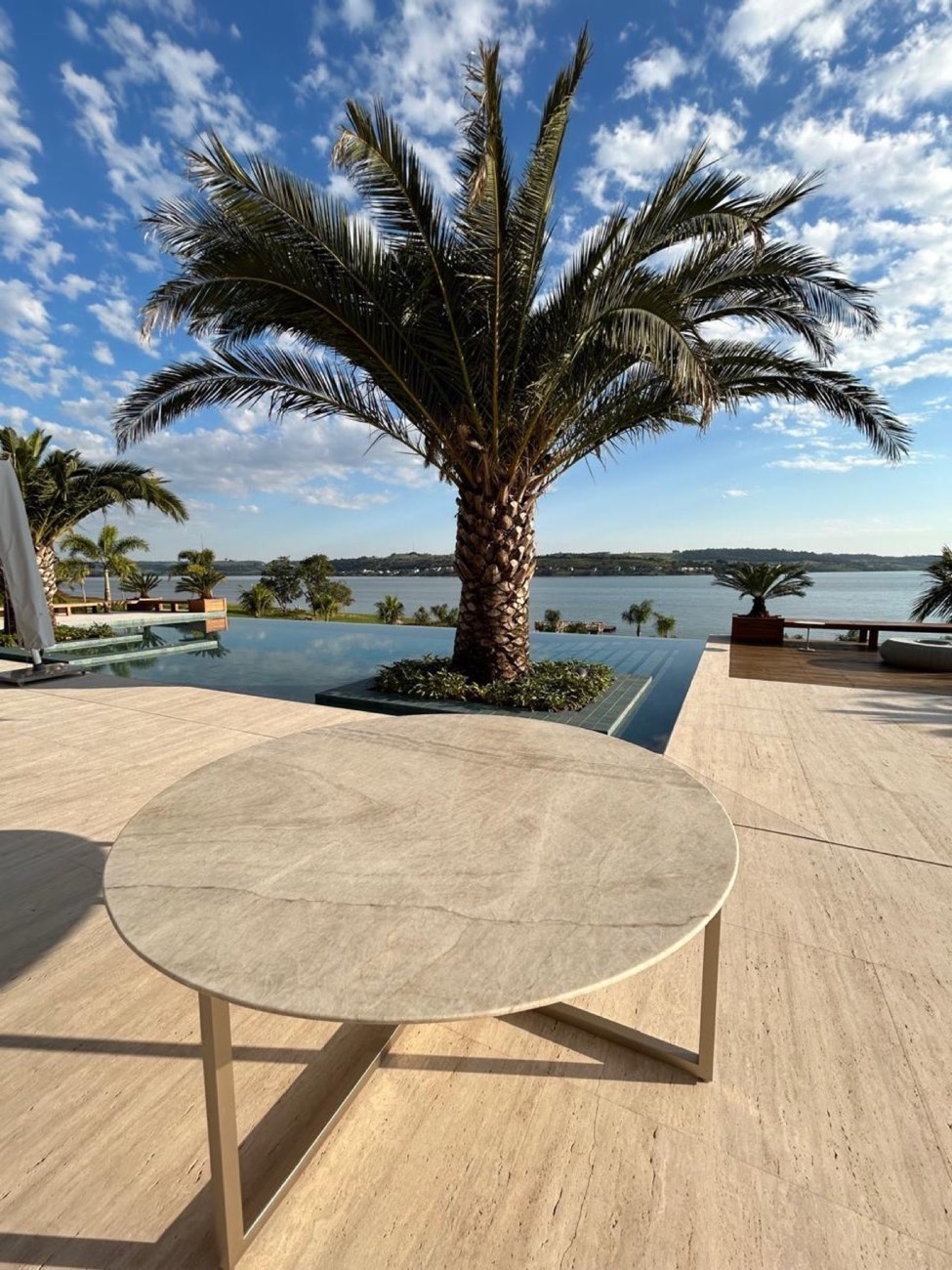 Deck externo em travertino claro com mesa redonda de pedra e vista para a água