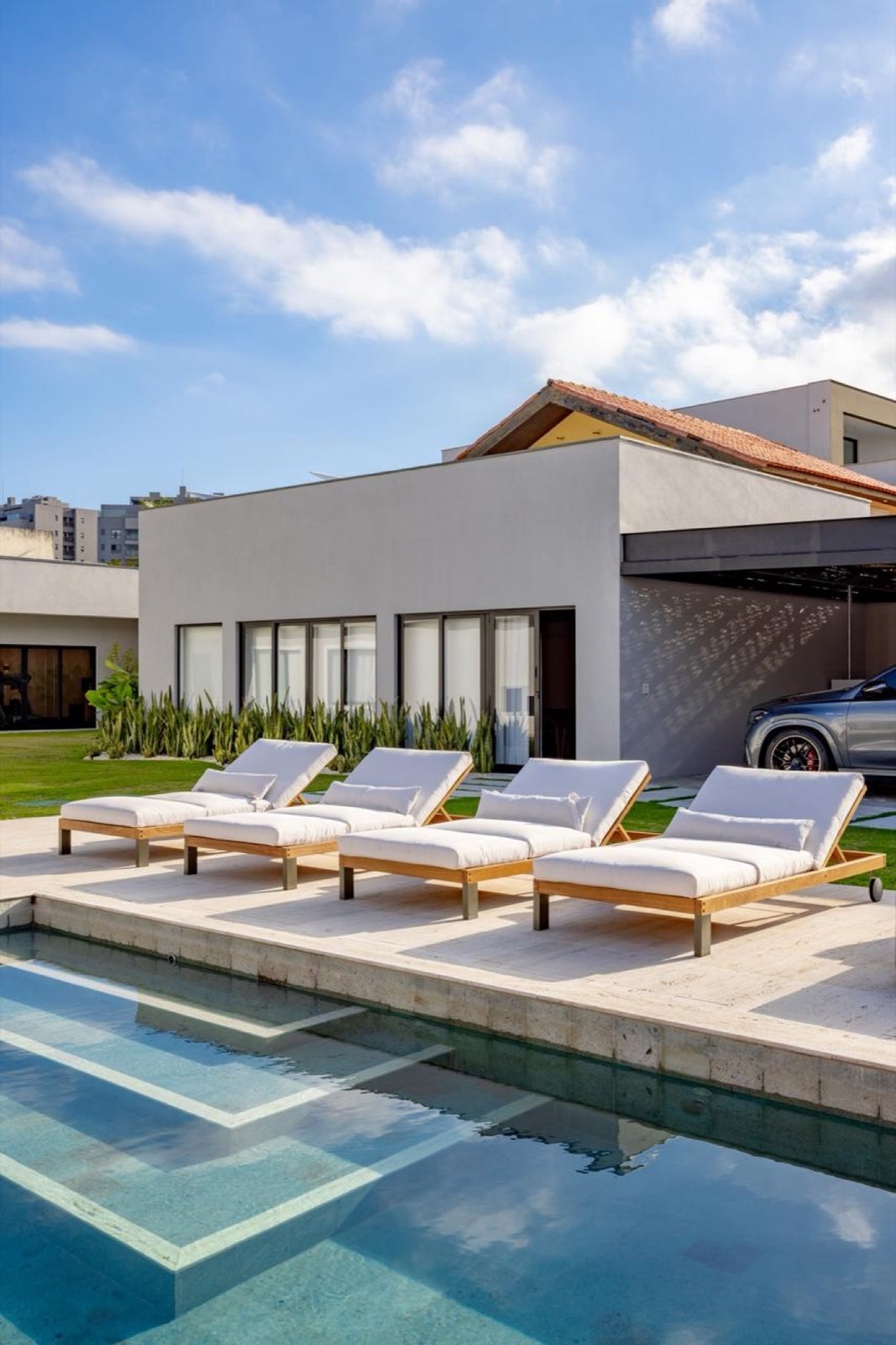Área de piscina com deck em travertino claro, espreguiçadeiras e casa moderna ao fundo